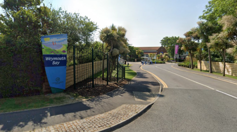 Entrance to Haven's Weymouth Bay Holiday Park. A sign with a painting of a beach on it is at the entrance. Behind it, a road leads onto the site, with palm trees either side. In the distance is a single-storey brick building.