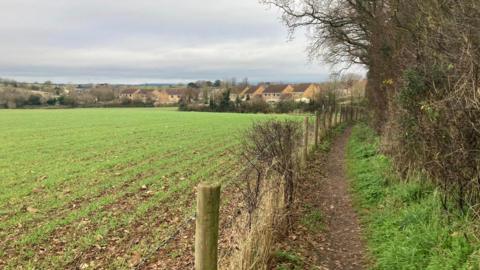 Walking path with a wire fence, green fields to the left and houses in the background 