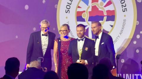 Broadcaster Jeremy Vine is pictured to the left of a group of four people on a stage. To his right is a blonde-haired woman in a dark red sequined dress and to her right is former Royal Marine commando Mark Ormrod MBE. Next to him is singer Matt Goss. All four are smiling and Mr Ormrod is holding an award.