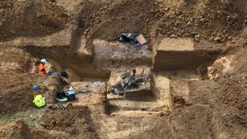 An archaeologist in an orange safety suit and white helmet working inside a deep trench, carefully excavating around a partially uncovered metal object, with tools and coloured markers scattered across the dig site.