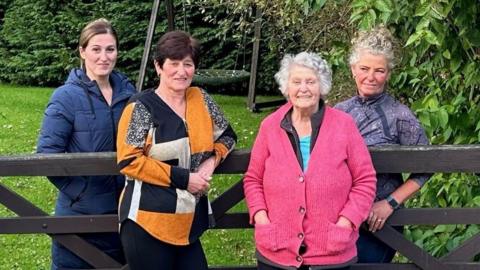 Kylie Cooper, Hayley Cooper, Angela Wiggans and Carol Tyrie standing outside smiling and leaning on a fence with trees and grass in the background