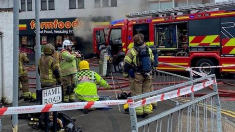 Several firefighters on a street corner in front of a fire engine which is in front of a Farmfoods store. Smoke billows from the front of the Farmfoods