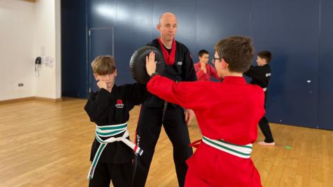 Two boys are practicing kickboxing in front of Matt. They are all wearing karate gis with one boy in a black gi and the other in a red gi. Matt is wearing a black gi and is looking at the boy in the red gi. The boy has thrown an open hand strike into a pad being held by the other boy. They are in a school hall with wooden flooring.