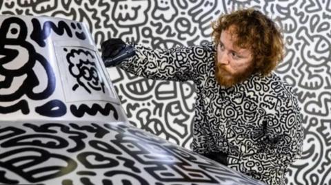 Mr Doodle to the right of screen wearing a shirt covered in his own doodles, with a doodle background and drawing on a structure in the foreground with a big black marker pen