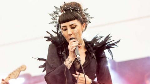 A female singer with brown hair, black clothes and an ornate metal tiara sings into a microphone