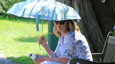 Woman sits under umbrella on a bench in the sun - holding a bottle of water