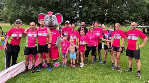 Parents and children who ran to raise money for a children's cancer charity. They are dressed in pink t-shirts with an elephant mascot in a park. 