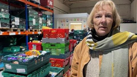 A woman in a coat, with a scarf around her neck is standing to the side of a large number of green crates, many of which have produce in them