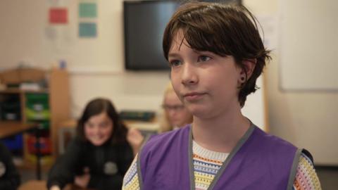 Mairia has short brown hair and is wearing a white striped jumper with a purple vest over the top