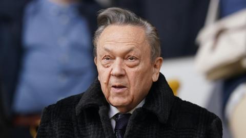 Former West Ham United chairman David Sullivan in the stands before the team's Premier League match at Molineux, Wolverhampton. He is wearing a black coat and tie with a white shirt.