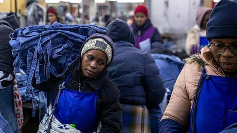 A woman, wearing a blue apron and black jersey, carries denim products in a garment factory in Lesotho