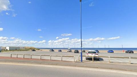 View of a seafront car park. It is a large open space, with a low warehouse type building and sand dunes to the left, and a road in front. There are a few cars parked and spaced out.  On the horizon are a number of off-shore wind turbines.
