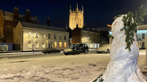 A snowman on Angel Hill in Bury St Edmunds, Suffolk. The snowman is near the camera on the right. In the background, snow blankets a car park which is empty apart from three cars. The picture is at night. The illuminated spire of a cathedral rises above houses on the other side of the car park.