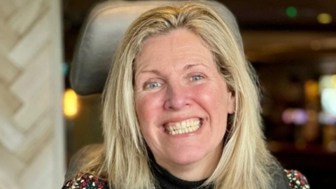 Nina Parry, who has straight blonde hair and is smiling widely at the camera. She is sitting in a wheelchair, with its headrest visible behind her. She appears to be in a pub or a restaurant, although the background is blurred.