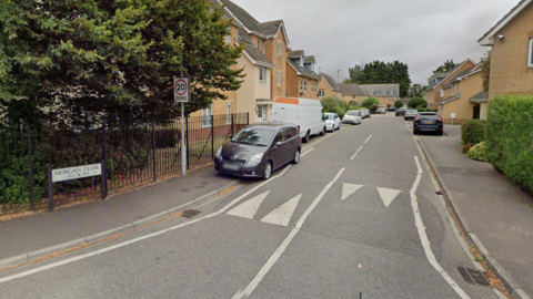 Morgan Close in Luton. The road is a residential area with houses on either side and is a 20mph zone which is signposted. Cars and vans are parked on either side of the pavement of the road.