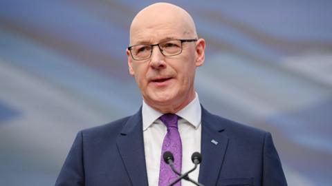 John Swinney, who is bald with glasses, speaks into a microphone. He is wearing a business suit and purple shirt 