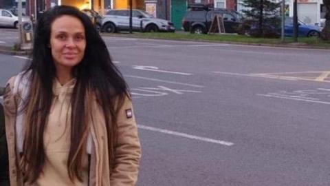 Jade Chapman, a woman who is standing outdoors, has long dark hair and is wearing a beige-coloured hoodie and coat. There is a road behind her and cars parked on the opposite side.