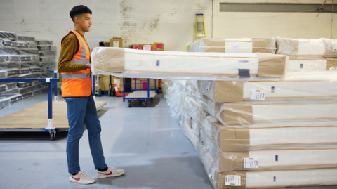A man in a hi-vis orange jacket, jeans and trousers is taking a wrapped mattresses off a stack. He is in a warehouse and there is a stack of mattresses to the side of him. 