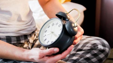 Hands holding an alarm clock. The person is wearing a white tshirt and black and white gingham pyjama bottoms.