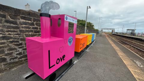 A brightly coloured pink train with 'Love' written on the side has pulled up on a small set of tracks on the platform next to a real railway line. The pink carriage has a chimney with a cardboard smoke plume emerging. It is pulling four carriages, one orange, one yellow, one blue and one green.