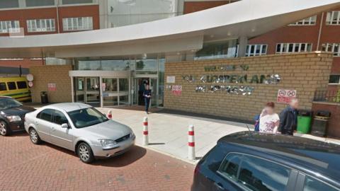 The main entrance at Cumberland Infirmary. A large sign on a brick wall reads "Welcome to the Cumberland Infirmary". Several cars are parked in front and people are entering and leaving the building. There is a curved, silver grey canopy above the entrance, behind which sits the modern red brick hospital building.