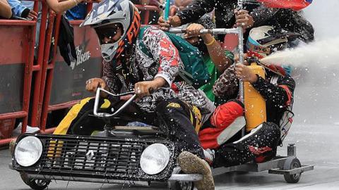 A soapbox cart on an urban course, with crew team members, one steering, one shooting a fire extinguisher and the third holding a flag