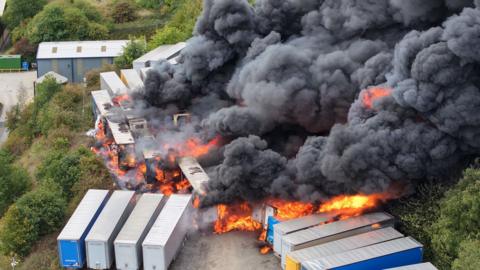 An HGV park on fire - at last 10 are on fire, with bright orange flames, and there is thick black rising smoke from the blaze above trees.