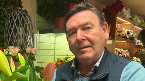 Michael O'Neill is holding a large bouquet of flowers. He is wearing a navy gilet and green fleece, dark hair and is staring in to the camera