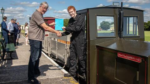 Matthew Forman-Clark, wearing a black boiler suit, stands on the step of a narrow-gauge diesel train receiving a blue certificate from a trainer, who is standing on the platform