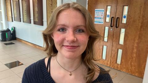 Kaitlyn is a teenage girl with long fair hair and blue eyes. She is wearing a dark blue v-neck top and is looking at the camera. 