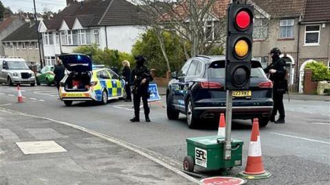 Armed officers can be seen stood next to a black car on a residential road. Another police car is parked to the left and has its boot door open. There is a temporary traffic light in the foreground.