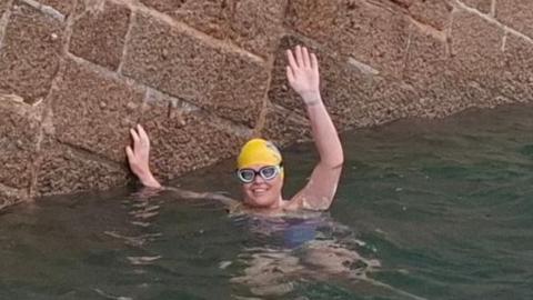 Marie-Helene Sanderson in the water. She is wearing goggles and a yellow swim hat and is waving at the camera while smiling broadly. Her other hand is on a stone sea wall.