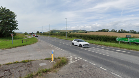 An photo taken from Google streetview. A white car sits on the central reservation at the Firle Road junction of the A27 between Beddingham and Polegate. The road is clear.