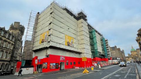 A general view of the G&V Hotel on George IV Bridge in Edinburgh. The hotel is covered by scaffolding with a white sheet over it. Green netting covers another part of the scaffolding. A red covered walkway goes around the bottom. The road is in the foreground.