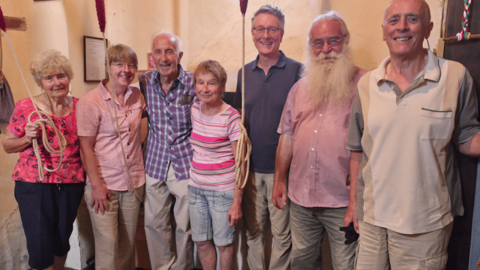 Seven men and women, all middle-aged or older, in a church tower, some holding bell ropes, and smiling to camera. 
