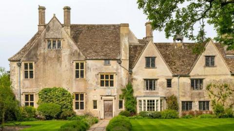 The front of a 16th Century manor house - lots of pitched roofs, stone window frames and chimneys. A large neat lawn and pathway out front, with tree branches hanging over from the right