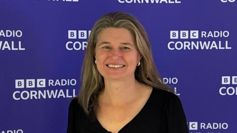A man with long light brown hair wearing a black v neck top standing in front of a purple backdrop dotted with the BBC Radio Cornwall in white letters. 