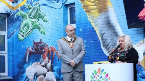Wolverhampton mayor wearing his chains of office stands besides a member of staff at the revamped garden at New Cross Hospital in Wolverhampton. She is standing at a podium and speaking into a microphone