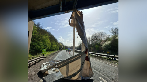A large piece of white material which has been torn off the side of a lorry hanging down from an overbridge on the A14. A roads policing officer is on the left of the image on the grass verge.