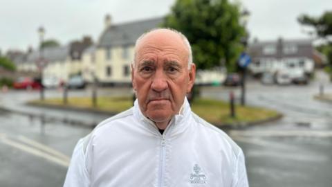Lee Newbon standing near the main square of the village with a little patch of grass and a tree behind him which is surrounded by roads. Behind that is his pub The Lamb. He is wearing a white sports jacket and has grey hair. 
