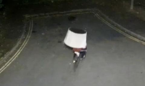 A man riding a bicycle whilst balancing a large white object on his head. It is unclear if the object is a lampshade or an upturned chair.