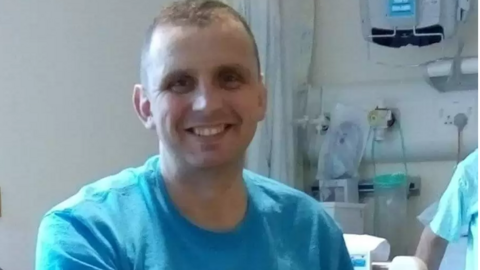 A smiling Rob Hope wearing a blue top is sitting in a wheelchair in a hospital room 