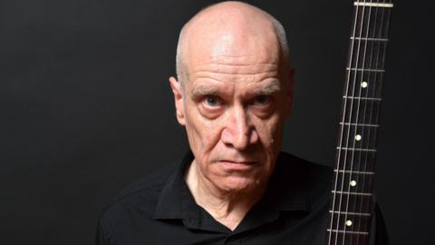 Wilko Johnson, who has blue eyes, stares directly into the camera with a serious expression on his face. He wears a black shirt and is pictured against a dark background. He's holding a guitar, where only the neck and fretboard is pictured.