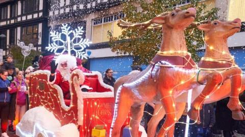 A Santa sleight drawn by two reindeers, with lights round it, going through a town. Lots of people are surrounding the display, with Santa Clause sitting in the middle, waving, wearing a red suit. There are toys on the front and shops decorated around. 