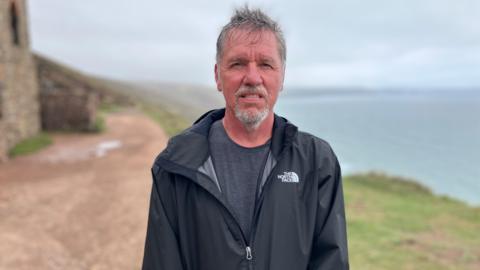 Richard Johnston smiling at the camera and wearing a black coat with a grey t-shirt. He is stood on a coastal path which has a sea view. 