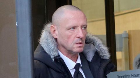 Mark Byrne is pictured looking off to one side in front of a court house. He is bald and clean shaven and wears a dark suit with a black parka on top with a fur-lined hood.