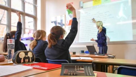 Classroom with three girls in uniform watching a teacher at a whiteboard. Two have their hands up.