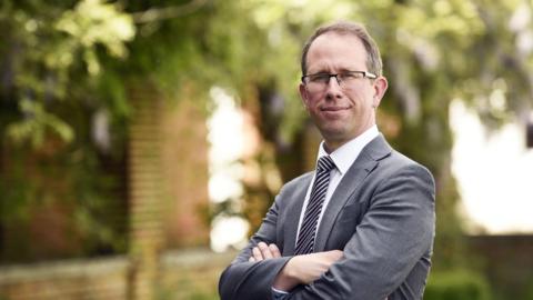 Matthew Barber, who has dark rimmed glasses, wearing a grey suit with his arms folded 