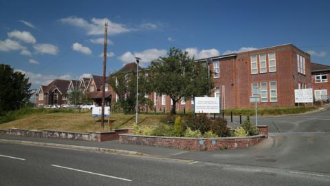 Llandrindod Wells Memorial Hospital, on a sunny day. The building itself is a red brick building which is two floors high. There are trees and a grass verge in front of the building, and there is a main road running alongside the hospital.