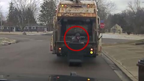 Man is hiding in a trash can with a garbage truck behind him.